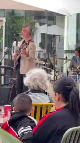 A band, including a singer playing bass guitar and a drummer, performs for an audience in an outdoor setting. The singer, with grey hair and glasses, wears a light tan jacket and black pants.  Behind the musicians are large glass windows reflecting the scene.  In the foreground, a few audience members are visible. A child in a black and red jacket is held by an adult in a red and black jacket. Another audience member with grey, curly hair sits in a yellow chair.  The scene is partially framed by green foliage on the lower left.