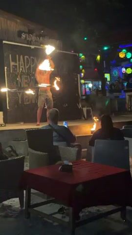 At night, an outdoor scene shows a fire performer on a raised stage with a "HAPPY NEW YEAR" sign in the background. The shirtless performer, wearing light-colored shorts, is actively twirling multiple flaming sticks. Two small flames are also visible on the stage floor. Below the stage, two individuals are seated at a table, facing the performer, with their backs to the camera. In the background, a bar area is brightly lit with numerous colorful string lights and spherical lanterns, where several other people are visible standing and seated. A table with a red tablecloth is prominent in the foreground. The ground appears to be sand.