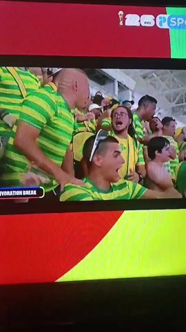 The video shows a screen displaying a soccer match. In the foreground, a man wearing a blue and white checkered shirt looks to his left with his mouth slightly open. Behind him, another person with short brown hair is partially visible. To the left of these men, a group of fans in green and yellow striped shirts are visible. The word HYDRATION BREAK is displayed in a small box on the left side of the screen. The background appears to be a stadium with blurred figures in the stands. The screen has a red and yellow border at the top and bottom.