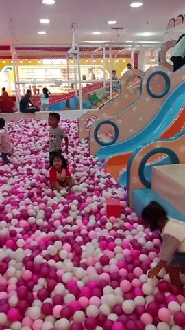 The video shows an indoor playground filled with pink and white ball pit balls.  Several children are playing in the ball pit, and two adults are also present, apparently supervising. The playground features a light-blue slide, a small, light-pink two-story structure, and other play equipment. The entire scene is brightly lit.  The camera moves slightly, shifting the view of the playground as it records. One woman is wearing a hijab.