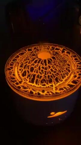 A small, round, white diffuser with an orange, cracked-earth design on top is the main focus of the video. The diffuser is emitting a light mist from the top. The light inside shines through the design, creating a lava-like effect. At the base of the diffuser, there is a small, lighted design of a sun and clouds. The background is dark and shows parts of a dark blue chair. The camera remains static and close to the diffuser throughout the short video. Music plays throughout.