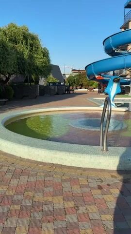 The video pans across an outdoor water park. In the foreground, a circular pool with green water is visible, bordered by mosaic tiles. Next to it, a larger pool with clear water is also visible. Two metal ladders lead into the pool. A blue water slide with a dolphin decoration winds down from a platform. In the background, there are buildings, trees, and lounge chairs. Two people are seated near the edge of the larger pool, and a third person stands nearby. The sky is clear and blue, suggesting it is a sunny day.