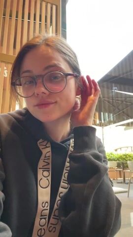 A young woman is seated, smiling, and waving towards the camera. She is wearing glasses and a black Calvin Klein hoodie. The background shows a partial view of an outdoor area with plants and architecture, likely viewed through a window. The indoor setting appears to be a cafe or restaurant.