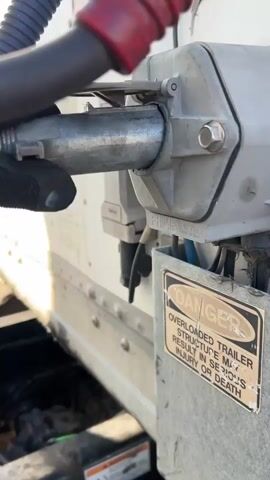 This video shows a close-up of the electrical and air connections on the rear of a semi-trailer. The scene appears to be outdoors, possibly a truck stop or loading dock, given the lighting and industrial setting. The sun is casting shadows, suggesting it's daytime, and the weather appears to be fair.

The primary objects in focus are the trailer's electrical and air brake hookups. A gloved hand is seen manipulating a metal connector, likely in the process of connecting or disconnecting it. Visible text on a warning sticker reads: "DANGER OVERLOADED TRAILER STRUCTURE MAY RESULT IN SERIOUS INJURY OR DEATH." Another label on the electrical junction box indicates "PHILLIPS 16-774 MADE IN MEXICO." A QR code is also affixed to the trailer's surface. The overall impression is one of routine maintenance or preparation for transport.