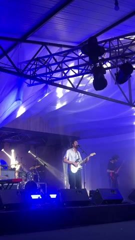 A band is performing on a stage, bathed in blue and white lights. A male vocalist in a light-colored shirt and dark pants stands center stage, playing a white electric guitar and singing into a microphone. To his right, another male guitarist in a dark shirt plays a dark electric guitar. In the background, a drummer is seated behind a drum kit. Several speakers are positioned on the stage. The stage is set against a dark backdrop, with a fabric ceiling overhead. The overall atmosphere suggests an indoor concert or performance.