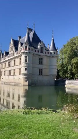 This video shows a large, ornate castle with a moat. The castle is light-colored stone with dark-colored roofs and spires. It has many windows and is reflected in the still water of the moat. There are green trees on the right side of the frame and green grass in the foreground. The sky is a clear blue. There are a few people visible on a walkway in the background to the right of the castle.