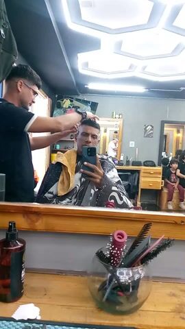 The video depicts a man getting his hair cut at a barber shop. He sits in a barber chair, draped with a black and white patterned cape, and is holding his phone up to take a selfie. The barber, wearing glasses and a black t-shirt, stands behind him, working on the man’s hair with clippers. A towel rests on the man’s shoulders. The scene is viewed through a large mirror reflecting the activity in the shop. 

In the foreground, on the counter in front of the mirror, there is a bottle of hair product and a round glass container filled with combs and hairbrushes. In the background, another barber works on a client in a different chair, and a child sits on a wooden ledge. The lighting in the shop is bright, with an interesting modern ceiling light fixture. A TV is mounted on the wall behind the barbers. Loud music is playing in the background.