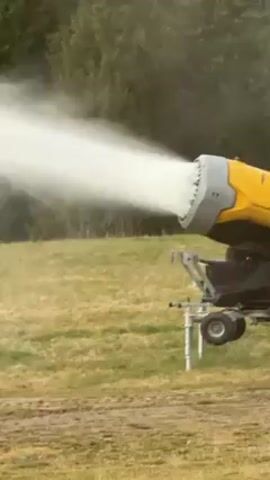 A snow cannon sits on a grassy field and shoots a wide stream of mist out into the landscape. The mist obscures the background until only a patch of the field is visible. The snow cannon is yellow and gray. There is a treeline in the background.