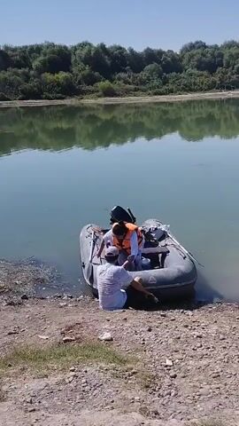 A small inflatable boat with an outboard motor is partially submerged at the edge of a calm lake. Two individuals are in the boat, one wearing an orange life vest. A second person is partially visible on the shore, near the boat, appearing to be interacting with the person in the boat. The surrounding environment consists of a rocky shoreline, with vegetation visible on the opposite bank of the lake. The sky is clear and blue.