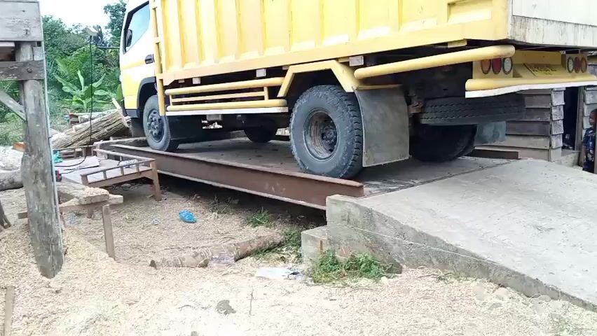 A yellow truck is positioned on a weighbridge. The truck's rear wheels are on the weighbridge, while the front wheels are on the concrete ramp leading up to it. A person wearing a yellow shirt and blue shorts is standing to the right of the truck, near a wooden building. Another person, partially visible, is also standing near the building. The ground around the weighbridge is covered with sawdust and some logs. The environment appears to be an outdoor industrial or processing area.