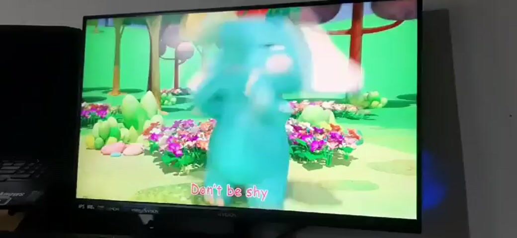 A television screen displays a colorful animated scene featuring a large, friendly blue elephant. The elephant is sitting down, holding a white tissue in its trunk. In front of the elephant, a smaller purple elephant lies on the ground, looking up at the larger elephant. The background is a bright green grassy field with scattered patches of colorful flowers in shades of pink, purple, and yellow. In the distance, there are stylized trees with red and green foliage. The scene appears to be set during the daytime in a cheerful, cartoonish outdoor environment.

Visible text on the screen reads: "When you really, really, really wanna sneeze". Below this, another line of text appears: "Wipe your nose". The animation seems to be part of a children's educational program or song, likely teaching about hygiene or how to handle a sneeze. The overall mood is happy and playful. The image quality suggests it is a recording of a screen, possibly taken in a home environment in Kawit, Philippines, given the provided location context. There are no distinct weather cues, but the bright lighting implies a clear day. The television itself is visible at the bottom of the frame, with a black bezel and a stand.