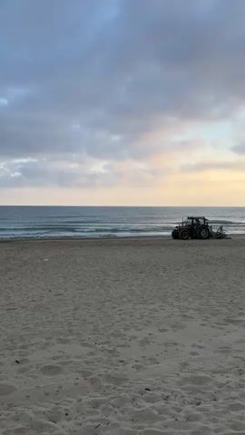 A tractor with a green attachment is parked on a sandy beach near the ocean. The water has gentle waves, and the sky is cloudy with hints of yellow and orange light. The sand is textured and appears undisturbed.