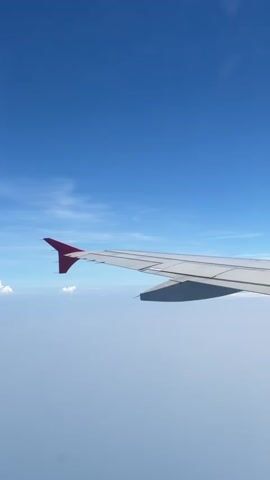 The video shows a view from an airplane window looking out at the sky and clouds. The wing of the airplane is visible in the foreground, with a dark shadow cast on the clouds below. The sky is a clear, bright blue with some wispy white clouds. The overall impression is one of a peaceful and scenic flight.