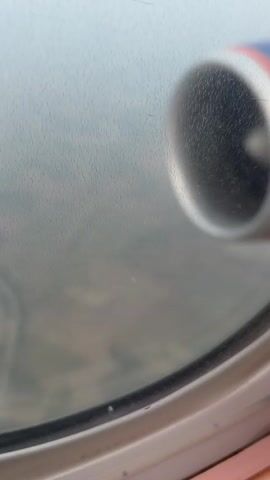 The video shows a view through an airplane window. The window is round and has a thick frame. The glass is slightly fogged and has some water droplets on it, suggesting that the plane is at a high altitude where condensation can occur. Outside the window, the landscape is visible but blurred due to the airplane's movement and altitude. The landscape appears to be a mix of green fields and possibly some structures like roads or buildings. A portion of the airplane's engine cowling is visible in the upper right corner of the frame. The engine cowling is metallic and has some colored accents. The overall impression is of a typical aerial view from a commercial airplane during flight.