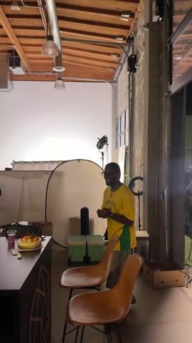 The video shows a group gathering in a studio space with a high ceiling featuring exposed wooden beams and industrial-style lighting fixtures. A man wearing a yellow t-shirt with "BRASIL" on it stands near a counter where a cake with lit candles sits. He appears to be leading the gathering and expressing his appreciation to the others, who are standing and sitting around a table. The space also contains a white backdrop, lighting equipment, and other studio-related items. The scene suggests a casual celebration or get-together in a creative workspace.