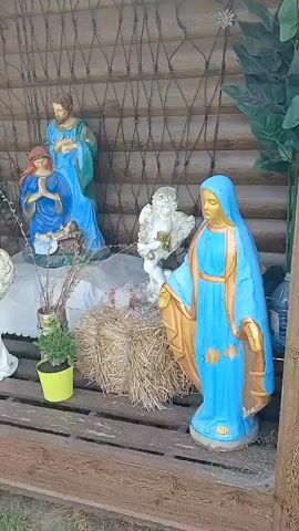 The video begins with a close-up shot of a nativity scene displayed in a wooden structure. Figures of Mary, Joseph, and an angel are visible, along with hay bales and potted plants. The camera then pans to the left, revealing more of the nativity scene, including a sheep figure and a decorative blue and yellow cylinder. The scene is set outdoors, with a grassy area and a wooden fence visible in the background. The video then transitions to an exterior shot of a building with bright yellow and blue walls. An arched window and a downspout are visible. The camera moves to the right, showing a gray asphalt ground surface. The video ends with a view of the side of the building.