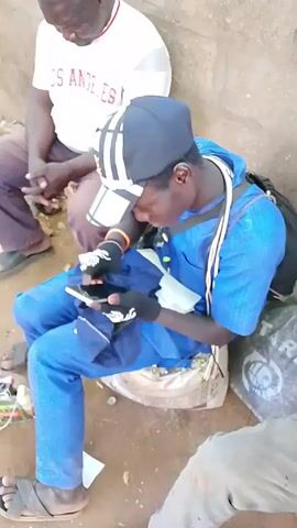The video shows three people sitting on the ground outside. A man in a white shirt that says “LOS ANGELES” sits with his hands folded in front of him. To the right of him sits a man in a blue shirt and pants and a black baseball cap. He is wearing black gloves with white graphics and working with what appears to be a mobile phone. There is a strap around his neck connected to some device clipped to his shirt. The third person is visible at the right edge of the video, but mostly out of frame. The person is wearing grey pants and brown sandals. The ground around them is dirt.