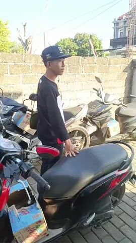 A man wearing a black long-sleeved shirt, a black baseball cap, and dark pants is standing next to a black motorcycle. He has a bag slung over his left shoulder. The man is looking to his right, away from the camera. The motorcycle is parked in an outdoor area with a brick wall in the background. There are other motorcycles parked around the man and the black motorcycle. The ground is paved with bricks. The sun is shining, casting shadows.
