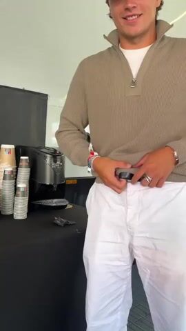 A young man wearing a tan quarter-zip sweater over a white t-shirt and white pants is standing in front of a black table. He is holding a black knife and appears to be adjusting his pants. To his left, on the table, are several stacks of paper cups and a coffee maker. The background is out of focus but appears to be a white tent or structure.