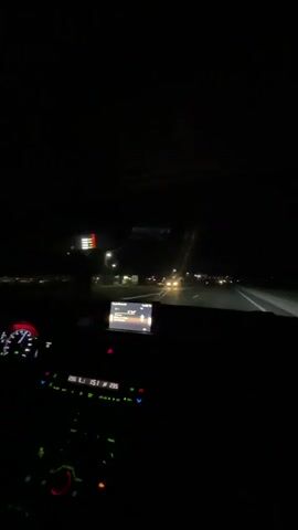 The video shows the interior of a car driving at night on a highway. The driver's side dashboard is visible on the left, with illuminated green and red buttons and a digital display showing numbers such as 23.5, 151, and 235. A car navigation screen is centrally located on the dashboard. The windshield shows the dark highway ahead, illuminated by distant streetlights and the headlights of other vehicles. The view is from the perspective of a passenger or the driver looking forward.