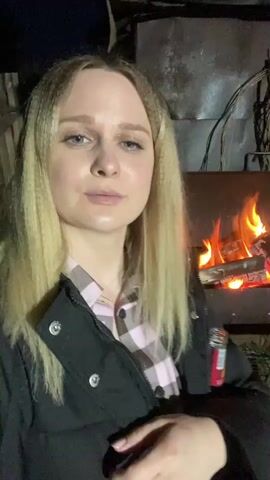 A young woman with blonde, crimped hair and blue eyes is featured in a close-up video, taken at night in Kauchuk Stantsiya, Russia. She is wearing a black puffer jacket over a pink and white plaid shirt. A small bottle with a red label, partially showing the text "VOSS", is held in her hand or jacket pocket. Behind her, a metal barbecue grill or brazier is filled with burning wood, producing bright orange and red flames that illuminate the background. Various metal structures and stacked wood logs are visible in the dimly lit outdoor setting. The woman maintains a calm, neutral expression as she looks directly into the camera. The scene suggests an informal outdoor gathering centered around a fire.