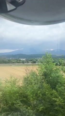 The video shows a view from a vehicle, likely a train or car, looking out at a rural landscape. In the foreground, there are green trees and bushes. Beyond that, a large, cultivated field stretches across the frame, with visible rows indicating it has been recently harvested or prepared for planting. In the middle ground, there is a collection of houses and buildings, suggesting a small town or village. Further in the distance, rolling hills or mountains form the horizon, under a cloudy sky. A speed limit sign showing "80" is visible in the middle right of the frame. The camera is moving, causing a slight blur to the scenery.