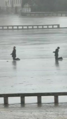 The video depicts a rain-soaked outdoor area. Two people are kneeling on the ground, seemingly searching for something. The person on the left is wearing a dark suit, and the person on the right is also dressed in dark clothing. A small white object is visible near the person on the left. In the background, there's a building and a barrier with horizontal bars. The rain is heavy, and the ground is covered in water.