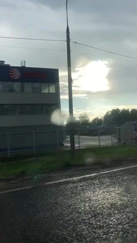 The video shows a view from a moving vehicle, looking towards a cloudy sky and a modern industrial building with blue and white accents. A fence runs along the front of the property, separating it from a road that appears wet, possibly from recent rain. A street lamp is visible, and in the distance, a gas station with covered pumps is seen. The sun is partially visible through the clouds, casting a bright glare. The video pans slightly to the right, revealing more of the building and the surrounding landscape, which includes trees and other structures. A white SUV is seen driving in the vicinity of the gas station. There are no people clearly visible or identifiable in the video.