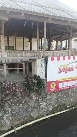 The video begins with a close-up of a banner advertising "WARUNG JEMBLEM Mak Yah" with a promotion offering discounts of 30%, 20%, and 10%. The banner is attached to a building with a rustic, open-air structure supported by wooden pillars. A small stream runs in front of the building, bordered by a stone wall. The camera then pans to the right, revealing a concrete path leading away from the building. The path curves and continues past a lush green rice field under a bright, clear sky. In the distance, there are trees and more buildings, suggesting a rural or semi-rural setting. The overall impression is of a simple, local establishment in a scenic, natural environment.