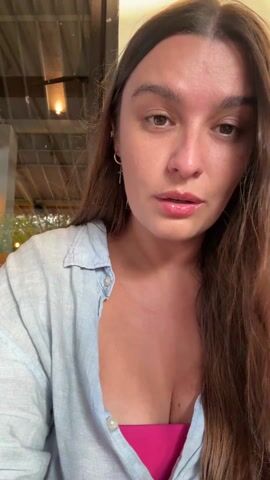A woman with long brown hair, wearing a light blue shirt over a bright pink top, looks directly at the camera as she holds a thin, metallic object with a green mouthpiece. She puts the mouthpiece to her lips and appears to make a "raspberry" or blowing sound. The background appears to be an indoor space with a view to the outside, possibly a cafe or restaurant setting. The ceiling has a light fixture visible.