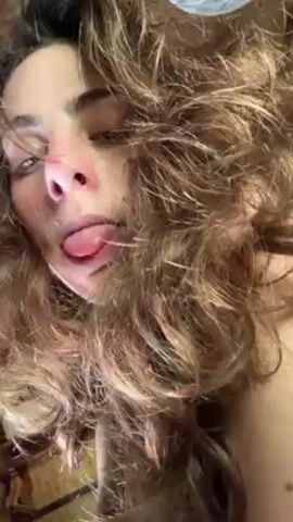 The video shows a woman with long, wavy brown hair lying on her back, looking directly at the camera. She extends her tongue out of her mouth, displaying it to the viewer. The background appears to be an interior space with indistinct objects. Her skin appears fair, and she seems to be indoors with soft lighting.