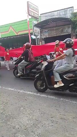A procession is moving down a street. Several people are holding a long red banner. Two people are riding on a black scooter, with the person in front wearing a helmet and the person behind wearing a red helmet. Two other people are on a red scooter further down the street. In the background, there are buildings with signs, one of which says "VINO PARTNERS" and another "TSAMARINA". The ground is asphalt and gravel.