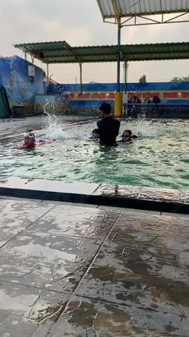 A group of children are swimming in a pool. A male instructor, wearing a black shirt, is supervising them. One child, wearing goggles and a pink swimsuit, is swimming in the foreground. Two other children, also wearing goggles, are in the middle of the pool with the instructor. Another child, wearing a red swimsuit, is visible in the background. The pool is surrounded by a tiled patio and a building with a covered roof. The sky is visible in the background.