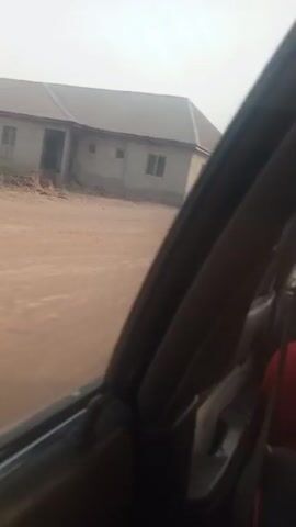 The video shows a view from inside a car driving on a dirt road. On the side of the road are two buildings that appear to be made of concrete or cinder block. The buildings have grey metal roofs and small windows. The sky is overcast.  The car is moving forward, but the road is bumpy, causing the camera to shake. The interior of the car is visible in the foreground, with a black window frame and a glimpse of a red seat. The audio includes someone speaking.