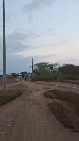 A dirt road stretches into the distance, flanked by dry vegetation and sparse trees. On the left side of the road, several buildings are visible. Two motorcycles are traveling on the road in the distance. A single utility pole stands tall on the right side of the road, with a wire extending upwards. The sky above is cloudy and appears to be during the twilight hours.