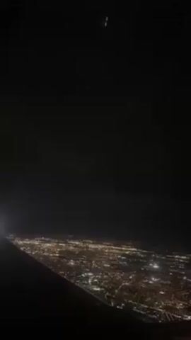 The view from an airplane at night shows the wing of the plane in the lower left and a large city sprawling below. The city lights twinkle in the darkness. The sky is almost completely black.