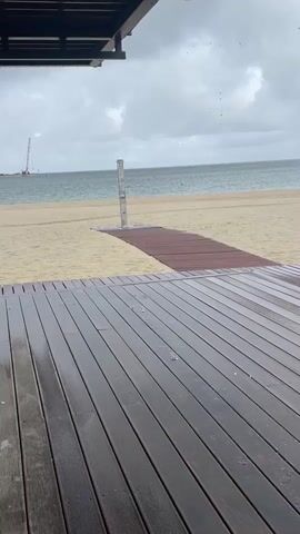 The video shows a view of a beach on a cloudy day. In the foreground, there is a dark wooden deck that appears wet, with water droplets visible. A wooden walkway leads from the deck towards the sandy beach. On the beach, there is a single shower stall with a silver pole. Beyond the beach is the ocean, and in the distance, a tall construction crane is visible against the horizon. The sky is overcast with dark clouds, and there is a light rain falling.