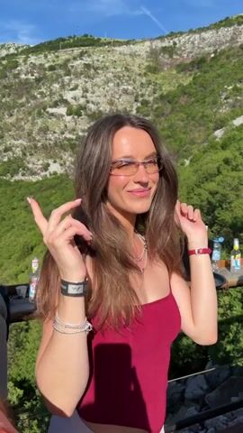 A young woman with long, brown hair and glasses stands on a balcony or patio overlooking a lush, green mountainous landscape under a clear blue sky. She is wearing a dark red, strapless top and light-colored pants. She is looking to her right and smiling. On a table behind her are bottles, an ice bucket, and other items, suggesting a gathering or a bar area. She moves her hands as she talks and looks around. The video transitions through slightly different angles and close-ups of the woman.