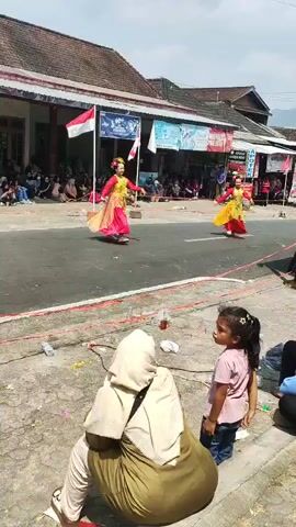 The video shows an outdoor street performance with two dancers in traditional attire. The dancers are wearing vibrant yellow and red outfits with decorative headpieces and are moving gracefully on the asphalt road. They are positioned in the middle of the road, with onlookers sitting on the side of the road observing the performance. Several flags, including what appears to be the Indonesian flag, are visible, suggesting a celebratory or cultural event. Buildings with tiled roofs line the street, and banners are displayed on them. In the foreground, a woman in a hijab and a young girl are seated on the ground, watching the performance. The ground is paved and slightly uneven, with some debris scattered around. The lighting suggests it is a bright, sunny day.