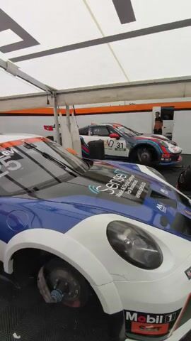 The video shows the inside of a garage or pit stop area, likely at a racetrack, with several race cars undergoing maintenance. The main focus is on a group of Porsche race cars, all with their wheels removed, revealing the brake rotors. The cars are mostly white with various color accents such as blue and orange. Mechanics and other personnel are seen walking around the vehicles. There are stacks of tires arranged along the back wall of the pit. The environment is well-lit with a white canopy overhead.  The floor is covered in a black and grey diamond pattern. Loud noises are audible, indicating the sounds of racing or maintenance.
