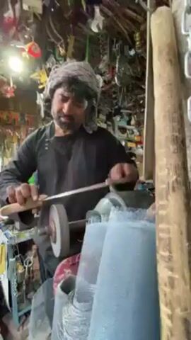 In an indoor workshop, a man with a beard and a turban-like head covering is actively sharpening a metal blade, likely a knife, using a bench grinding wheel. He is dressed in a dark long-sleeved garment. As the blade makes contact with the spinning wheel, a cascade of bright orange and yellow sparks erupts and flies outwards, illuminating the immediate area around the grinder. The man's gaze is directed downwards, focused on the task at hand, his expression neutral.

The background reveals a densely packed shop environment, characteristic of a traditional workshop or repair store in Kandahar, Afghanistan. Numerous tools, metal components, and various other objects are haphazardly hung on walls and shelves, creating a cluttered but functional space. A bright artificial light source is visible in the upper left corner, providing illumination to the scene. No discernible text is visible within the frame. The scene captures a moment of focused craftsmanship, highlighting the manual labor involved in sharpening tools.