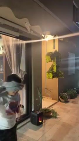 A person wearing a blindfold and holding a toy sword swings at a piñata shaped like the number "21". The piñata is gold, green and black. The person is wearing a white t-shirt, camouflage pants and a grey scarf around their head. The scene appears to be outside, with a patio and glass doors visible. A small black speaker sits on the ground near the piñata. There are plants in pots and a cabinet inside the house visible through the glass doors. Someone is laughing in the background.