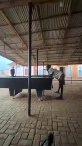 The video shows two people playing pool under a corrugated metal roof in an open-air structure with brick flooring. One person, wearing a white shirt and dark shorts, is leaning over the pool table taking a shot, while the other person, dressed in dark clothing, is standing at the opposite end of the table. The pool table is black with a green surface. The camera is stationary, providing a wide view of the scene. Outside the structure, there are brightly colored buildings and a patio area with red tables and chairs. The overall atmosphere is casual and relaxed.