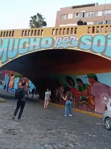 The video shows a street scene with colorful mural art on the walls and under a bridge.  People are seen walking and taking pictures of the artwork. A silver car and a blue car are parked on the street. In the background, a building with a domed tower is visible. The street is paved with cobblestones.  The phrase "Mucho por Sonar" is written on the underside of the bridge, and "Mucho por Hacer" is written on the wall.  A tree is in front of one section of the mural.