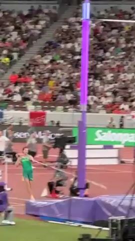 The video shows a male pole vaulter in a green and yellow uniform competing in front of a large crowd in a stadium. He successfully clears the bar, which then falls onto the crash pad. The vaulter lands safely on the crash pad as well, and quickly stands up to look at the result. A judge standing nearby observes. Banners are visible in the background, advertising Salonpas and Japan Airlines. A flag of Kiribati is also visible.