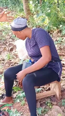 A woman is sitting on a wooden stool in a wooded area. She is wearing a gray head covering, a dark blue short-sleeved shirt, and black leggings.  She has a small black container and a larger, white, translucent bucket next to her.  She holds a small stick in her hand, looking away.  A man's arm and leg are visible at the bottom left corner of the frame. The ground is covered with leaves and small plants. Sunlight filters through the trees.