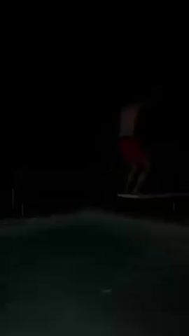 A person wearing red shorts stands at the end of a diving board at night, over a pool with greenish water, then jumps in. The person is only partially visible in the dark.