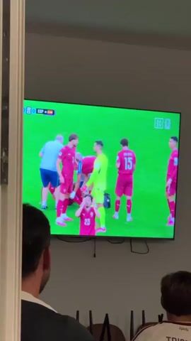 The video shows a group of people watching a soccer game on a large television screen. The game appears to be between Spain and another team wearing red uniforms with a crest that includes a shield and stars. On the screen, players in red uniforms are gathered around a fallen player. One player in a yellow goalkeeper's uniform is also present. A small child wearing headphones and a pacifier is visible in the foreground, sitting on someone's lap and holding a blue cup. A woman next to them is smiling and appears to be reacting to the game. The room is dimly lit, suggesting the game is being watched in the evening or in a darkened room. There are chairs visible in the lower part of the frame, indicating a viewing area.