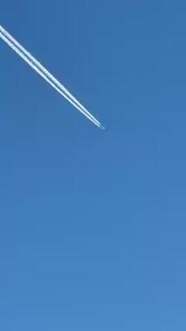 The video shows an airplane with two visible contrails in a clear blue sky, viewed from a ground perspective. The camera then pans down to reveal a landscape with bare trees, a house, and mountains covered with snow under the same clear blue sky.