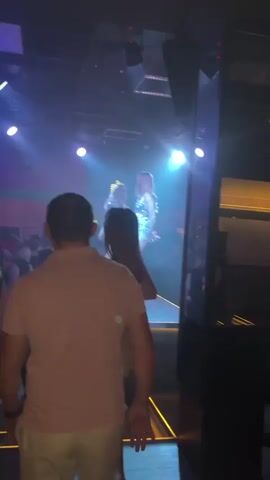 The video shows two distinct scenes in what appears to be a nightclub or bar. The first scene, at the beginning of the video, features a person on a stage under bright stage lighting, possibly performing. The audience is visible in the foreground, with a person in a pink shirt facing away from the camera. The second scene, which transitions in, shows a darker environment with blue LED lights on the ceiling and several people gathered. The camera is focused on the backs of several individuals.